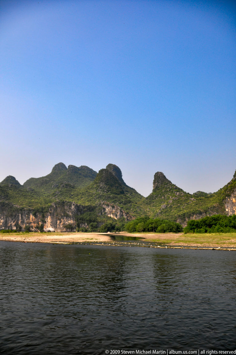 Wide bend of the Li River flowing beside continuous pale limestone cliffs in a tropical karst landscape, with exposed gravel bars along the banks.