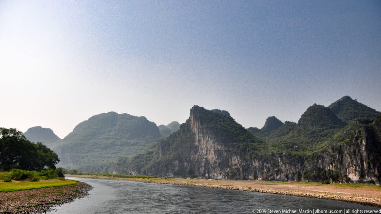 Li River bend with a pale gravel bar on the inner curve, steep vegetated karst cliffs on the opposite bank, and small boats moving through the channel.