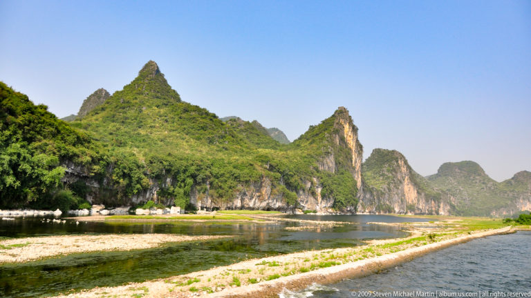 A bend of the Li River with a pale gravel bar on the inner curve and steep vegetated karst cliffs on the outer bank.