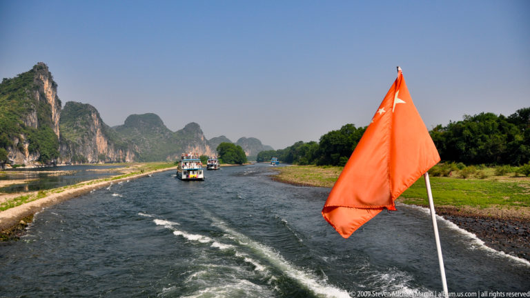 Orange flag above the V-shaped wake of a Li River cruise boat, with other passenger vessels and karst peaks in the distance.