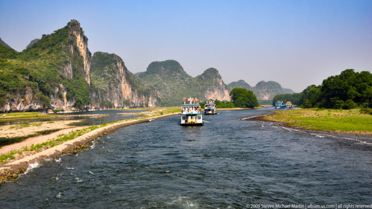 Several Li River tour boats travel in the same direction through a narrow karst river channel bordered by exposed gravel bars and high limestone cliffs.
