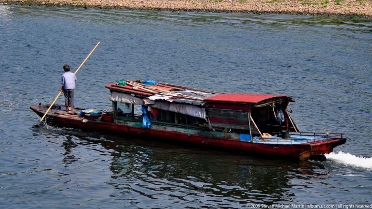 Boatman standing at the stern of a long covered wooden riverboat, propelling it with a bamboo pole on the Li River.