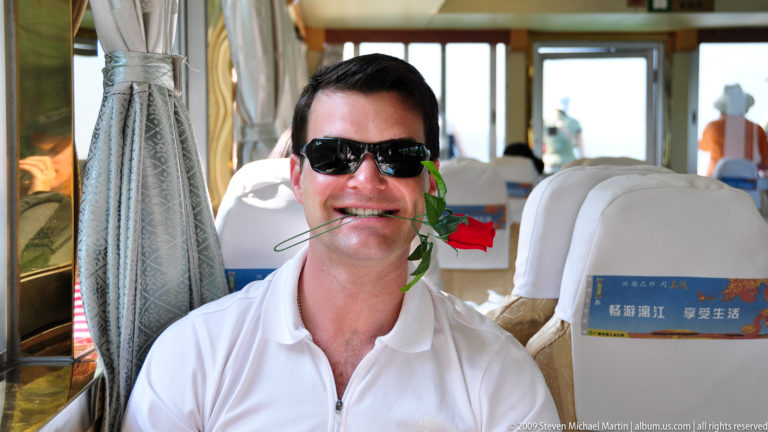 Man wearing sunglasses holding a red flower in his mouth inside a passenger boat cabin.