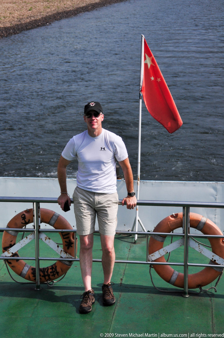 Man in a white athletic shirt standing by the stern rail of a moving Li River cruise boat, with two orange life rings on the railing and a red flag behind him over the river.