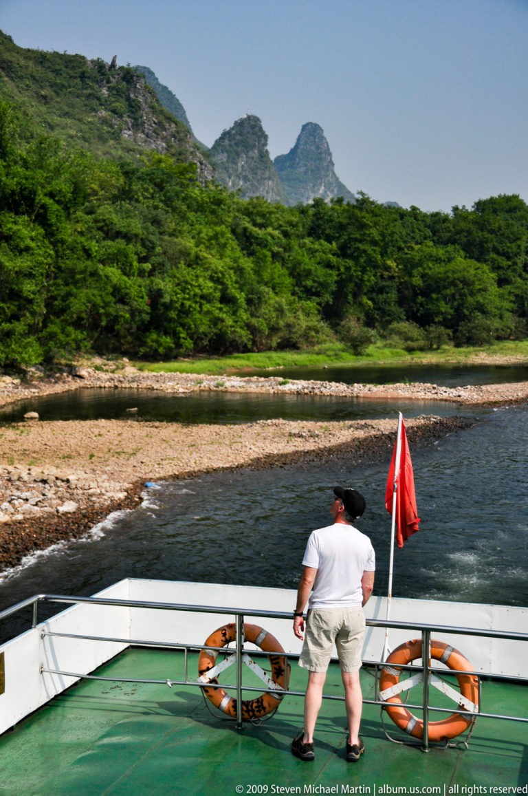 Rear view of a man on a Li River cruise boat looking toward a curved gravel bar and exposed riverbanks.