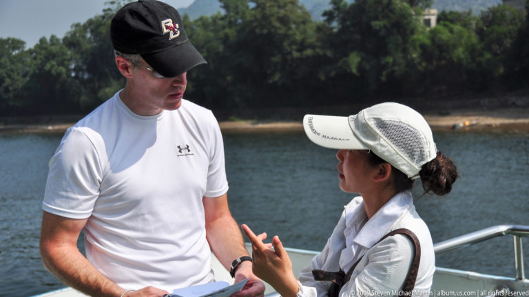 Male traveler speaking with a female tour guide on the outdoor deck of a boat with the Li River in the background.