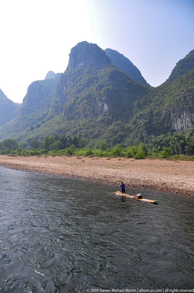 Person standing on a narrow bamboo raft, poling through shallow water beside a broad gravel bank and karst cliff.