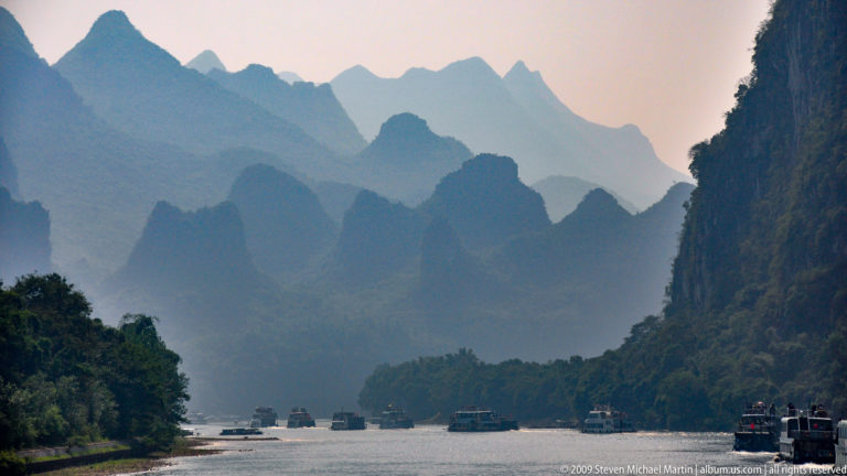 Li River corridor with layered hazy karst mountains and cruise boats on a broad channel framed by vegetated banks.