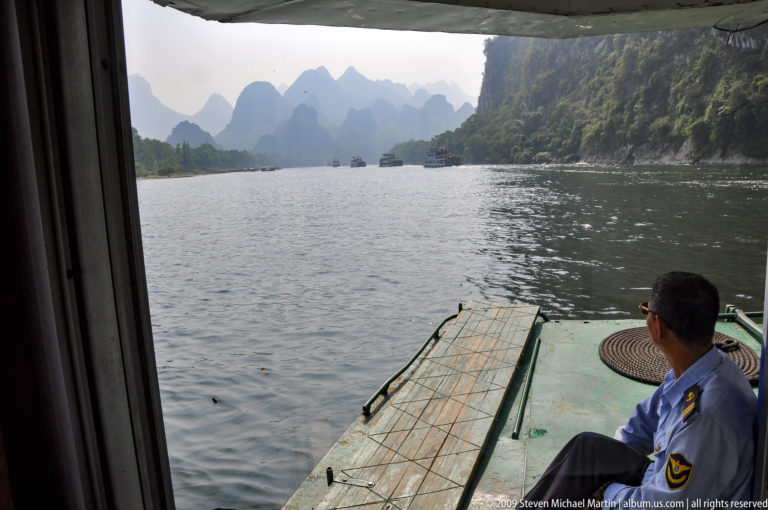Crew member in a blue uniform seated on the green deck of a working boat on the Li River, looking ahead toward karst cliffs and other vessels.
