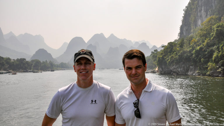 Two men standing on the deck of a Li River cruise boat with karst mountains and other boats behind them.