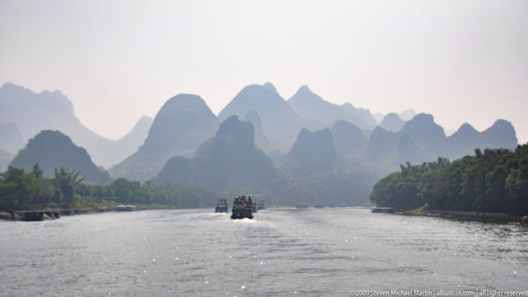 Layered hazy karst mountains rise above the Li River corridor with cruise boats on the water and vegetated banks on either side.