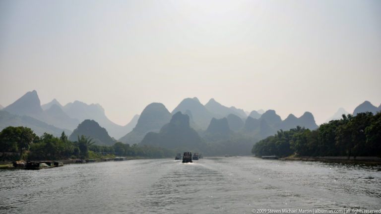 Li River in Guilin with cruise boats on a broad channel and hazy layers of blue-gray karst mountains in the distance.