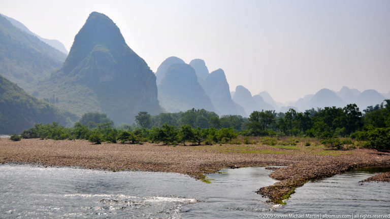 Shallow river channel splitting around a broad gravel bar with vegetated floodplain and hazy karst mountains in the background.