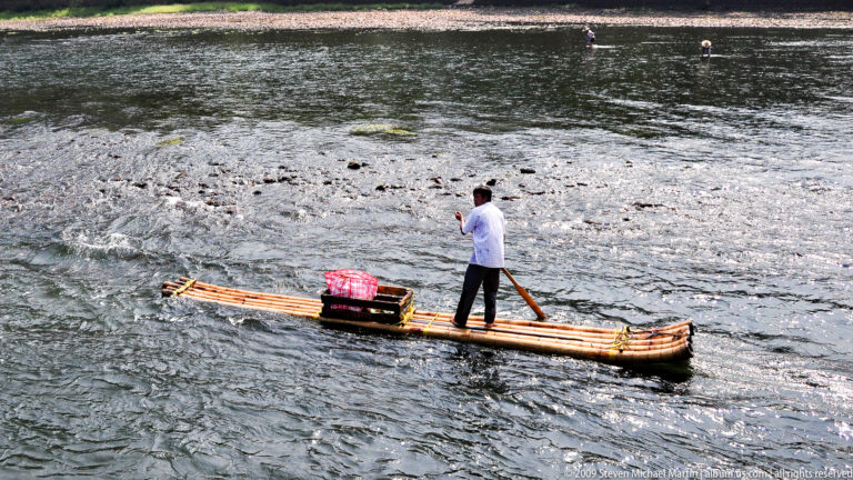 Man standing and poling a loaded bamboo raft with a pink-covered bundle and wooden crate through shallow, flowing river water.