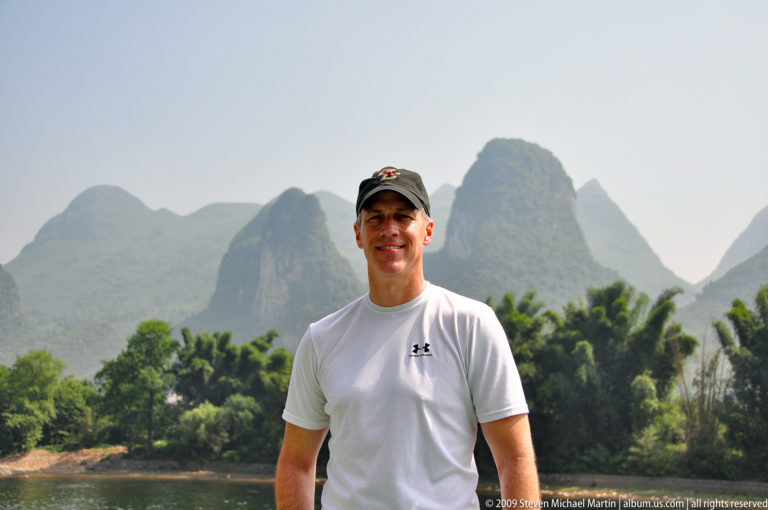 Man in a white athletic shirt standing by the Li River with karst mountains and dense palm-like vegetation in the background.