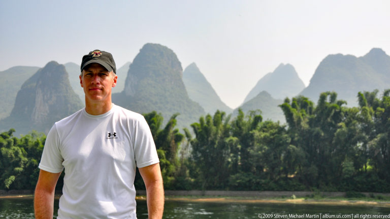 Man in a white athletic shirt posing by the Li River with karst mountains and dense palm-like vegetation in the background.
