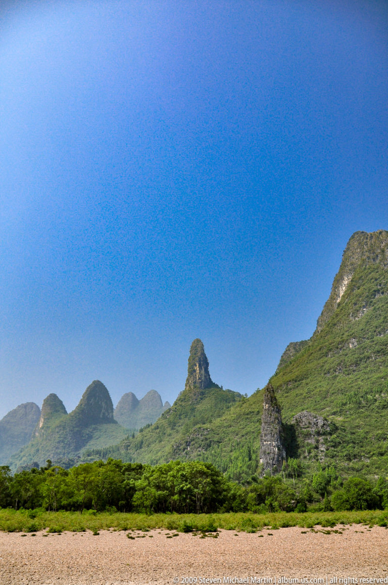 Slender limestone spire rising among broader hills under a deep blue sky in Guilin.