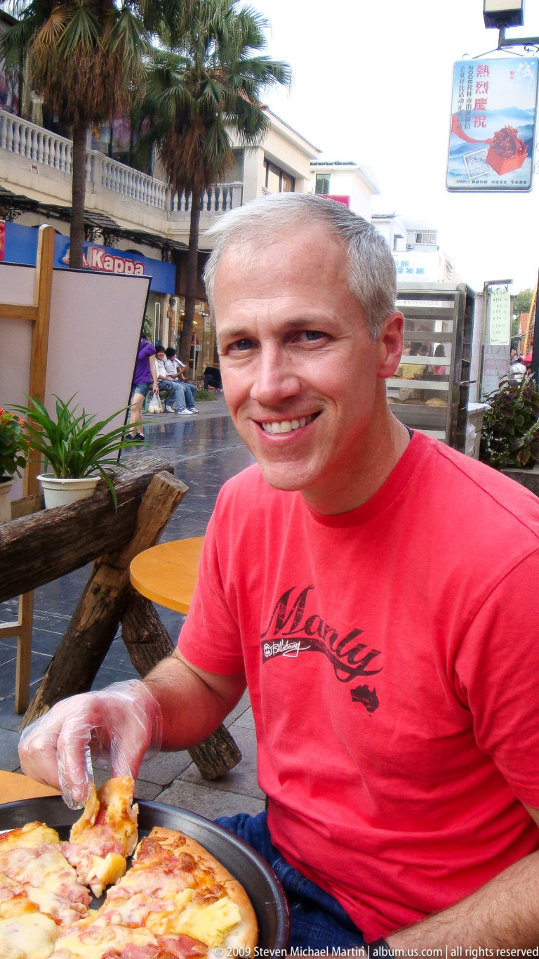 Gray-haired male traveler in a red T-shirt smiling at an outdoor Guilin café, shown from the chest up with blurred tables and plants behind him.