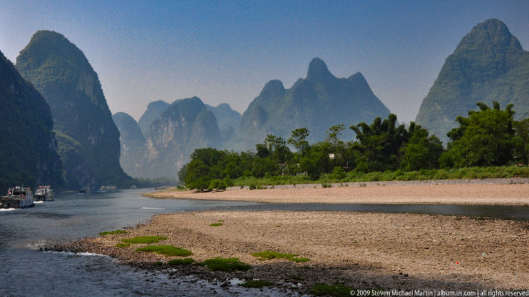 Wide gravel bar extending into the Li River with overlapping karst peaks in the distance.