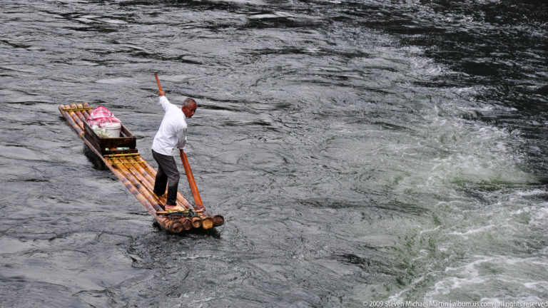 Man poling a low bamboo raft with a covered load through swirling river current on the Li River.