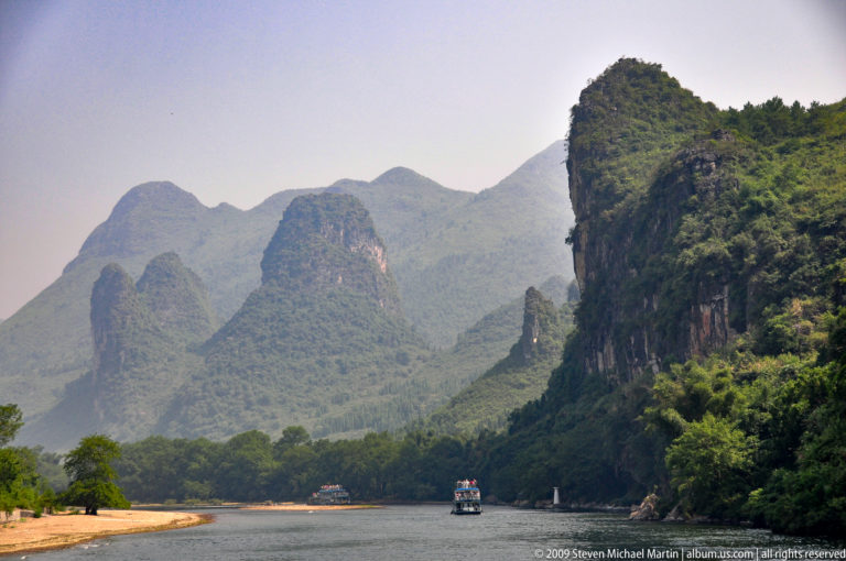 Cruise boat traveling through a narrow Li River gorge beneath a towering karst cliff.
