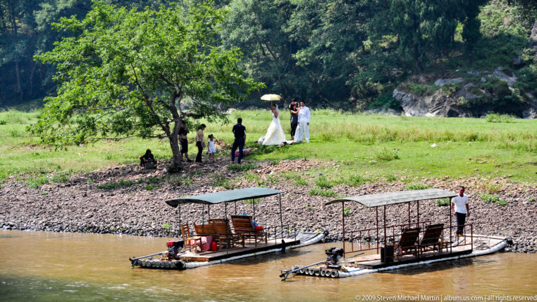 Bride in a white gown with a small photography group on a grassy Li River bank, with covered bamboo rafts floating in the foreground.
