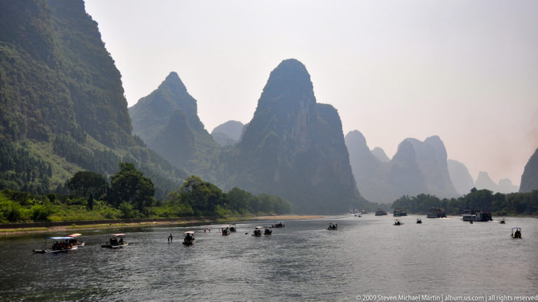 Many small boats and rafts scattered across the Li River beneath a tall, sharply pointed karst peak in Guilin.