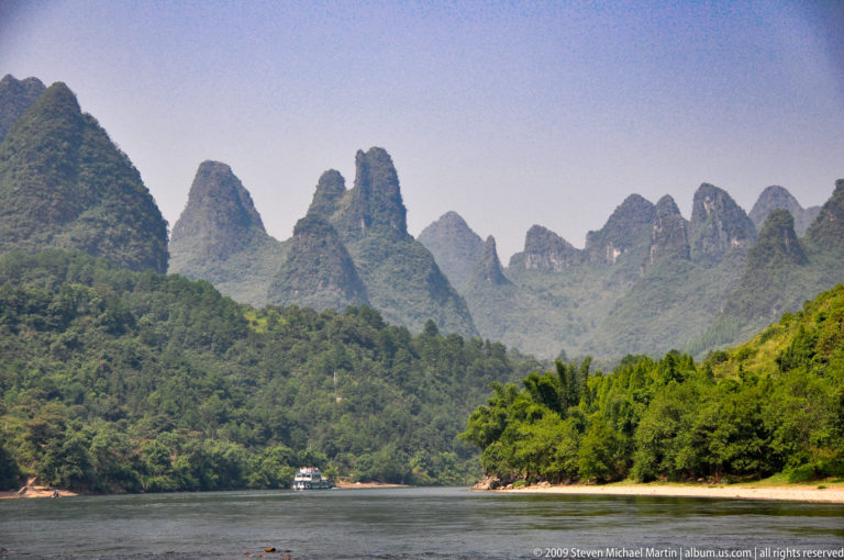 Calm Li River channel leading toward closely packed needle-like limestone peaks, with dense trees along the right bank and a small cruise boat in the distance.