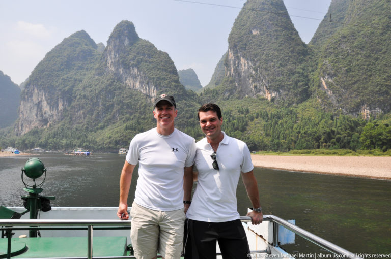 Two male travelers posing side by side on a boat with the Li River and Guilin karst mountains in the background.