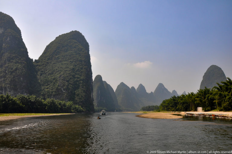 Boats travel along a narrow Li River channel in Guilin, hemmed in by steep rounded limestone karst towers and a sandy bank.
