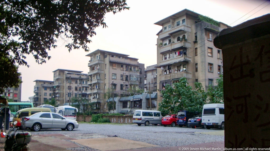 Weathered mid-rise Guilin apartment blocks with balconies and rooftop vegetation around an unpaved parking area with parked cars and vans.