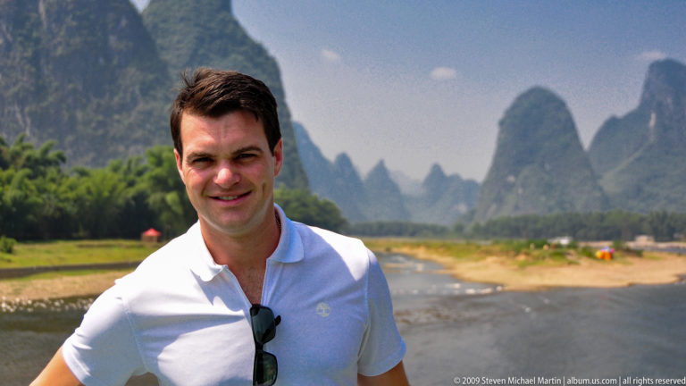 Smiling male traveler posed by the Li River with soft-focus karst peaks in the background.