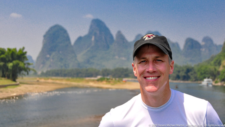 Smiling male traveler posed by the Li River with limestone karst peaks softly visible in the background.