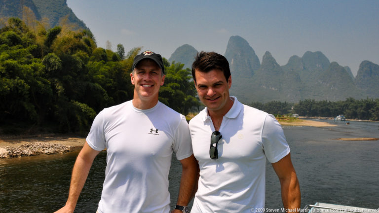 Two male travelers standing together on a boat with Li River and karst mountains in the background.