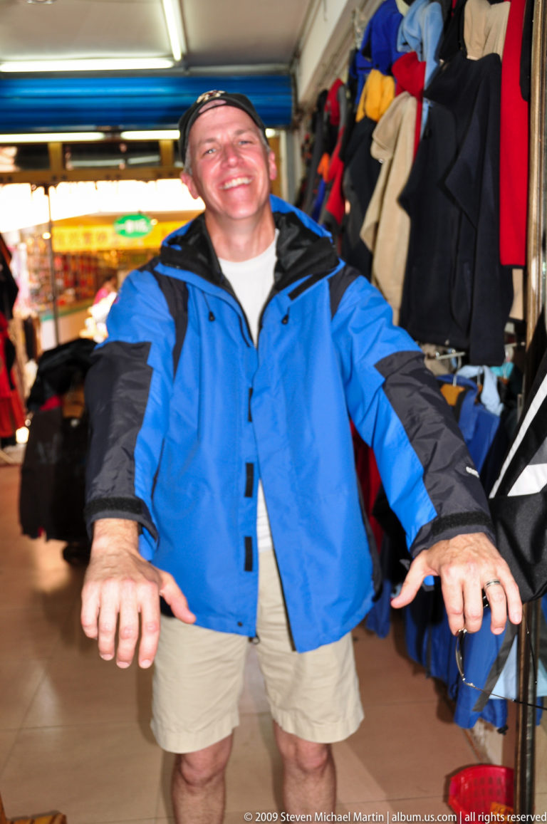 Torso and hands of a traveler trying on a bright blue outdoor jacket among clothing racks in a Guilin market.