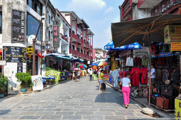 Stone-paved pedestrian shopping street in Yangshuo lined with colorful shopfronts, umbrellas, and cafés, with people walking between tourist-oriented stalls.