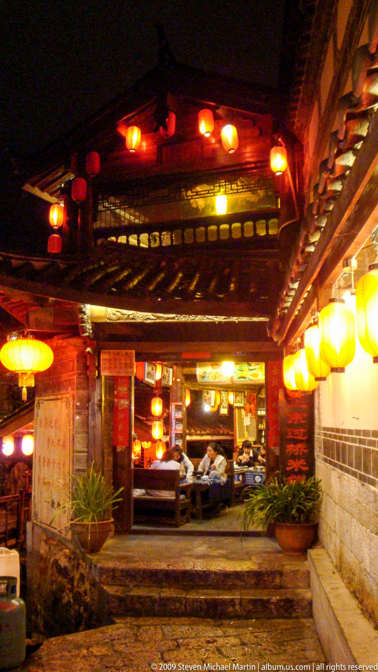Lantern-lit traditional timber restaurant passage with exposed beams, carved brackets, and seated diners in Lijiang Old Town, China.