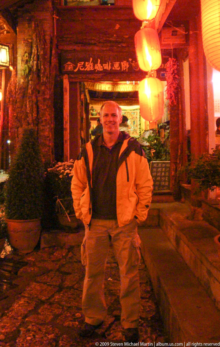 Male visitor in an orange jacket standing in a lantern-lit Old Town lane with potted flowers and traditional storefronts in Lijiang, Yunnan, China.