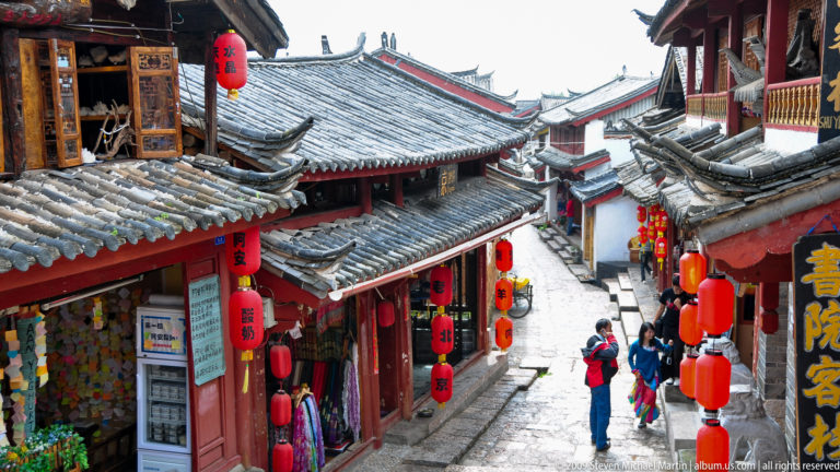 Elevated view down a narrow stone street in Lijiang, China, with red lanterns, overlapping grey-tiled roofs, and visitors walking along the sloping lane.