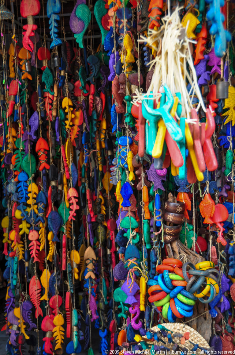 Dense hanging display of brightly colored fabric and plastic charms, including fish, dolls, tassels, and cords in turquoise, red, yellow, and blue, at a market in Lijiang, Yunnan, China.