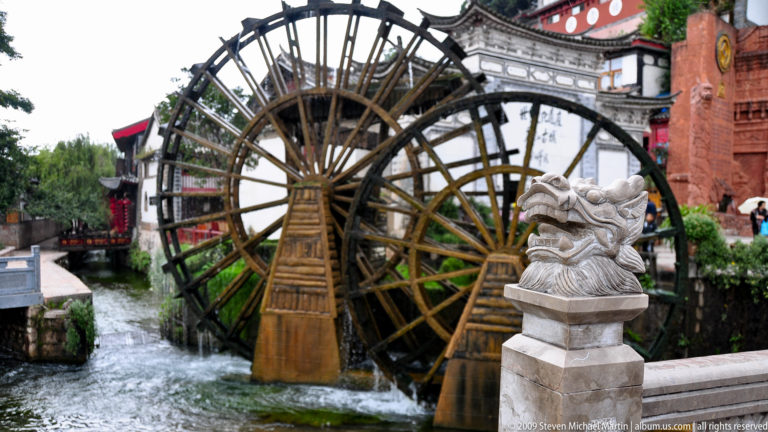 Two large wooden waterwheels in moving water beside a pale carved stone animal head and grey-tiled traditional buildings in Lijiang, Yunnan, China.