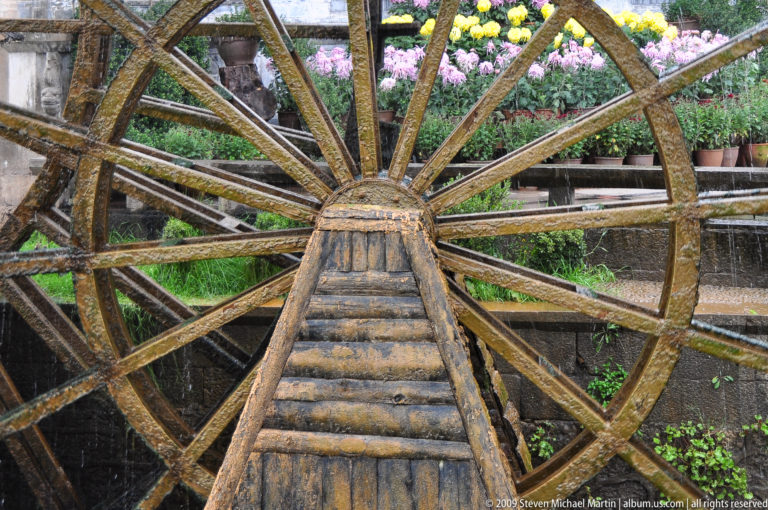 Close, front-on view of a traditional wooden waterwheel in Lijiang, Yunnan, China, showing its radial spokes, stepped central hub, dripping water, and potted flowers in the background.
