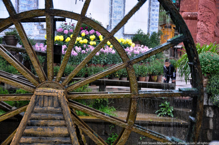 Close view of a large wooden waterwheel in Lijiang, Yunnan, China, with heavy spokes and water spilling from its paddles beside rows of flowers.