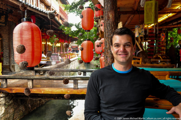 Male visitor smiling toward the camera while seated beside a lantern-lined canal with red lanterns, hanging pinecones, and flowing water in Lijiang, Yunnan, China.
