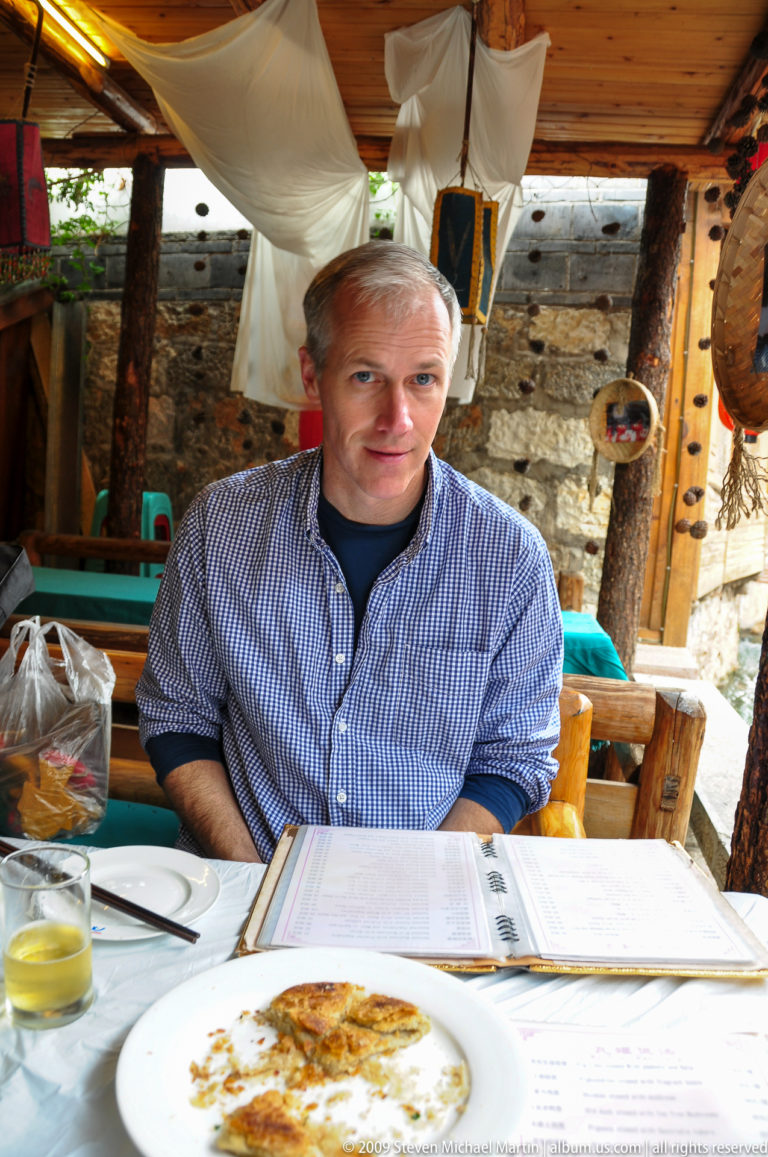 Male visitor in a blue checked shirt seated at a rustic wooden table against a stone wall with heavy timber furniture in a Lijiang restaurant, Yunnan, China.