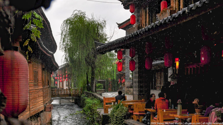 Rainy canal-side restaurant lane in Lijiang Old Town, with diners seated under red lanterns beside a rushing canal and dark timber buildings.