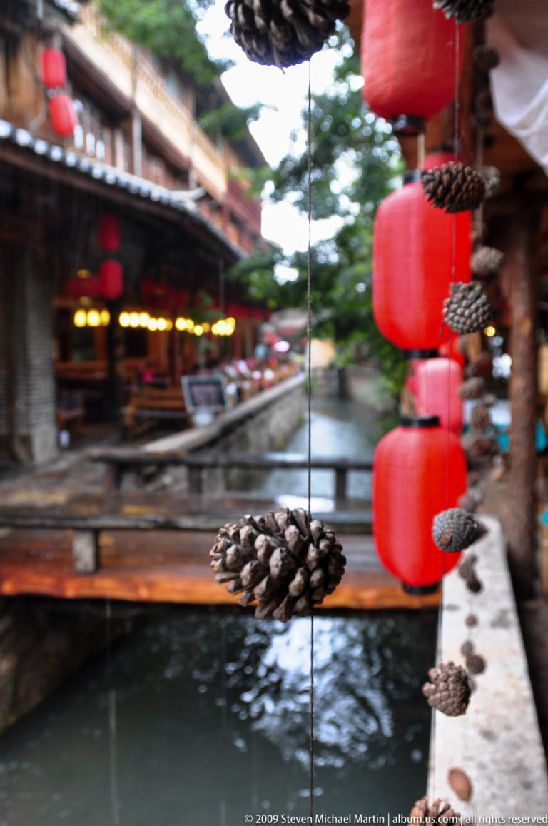 Single pinecone hanging from a thin cord with red lanterns and a narrow canal blurred in the background.