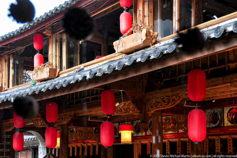 Traditional wooden building in Lijiang, China, with carved brackets, grey tile eaves, and rows of cylindrical red lanterns on both levels.