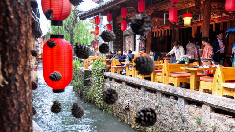 Traditional restaurant terrace beside a narrow canal in Lijiang, Yunnan, with red lanterns, hanging pinecones, and wooden tables above a stone wall next to flowing water.