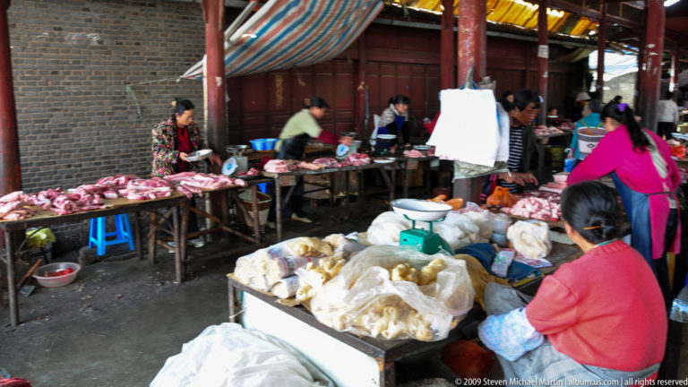 Indoor row of butchers in Lijiang, China, selling cuts of raw meat laid out on wooden tables under a striped canopy.